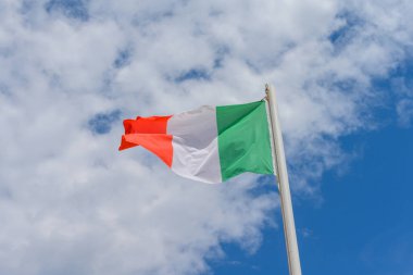 Italian flag waving in the wind. It is a beautiful sunny summer day, with blue sky and white clouds in the background.