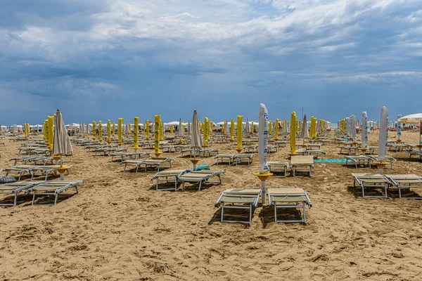 Beach in Caorle, Italy, with rows of sun beds and closed parasols. The beach is almost empty, except for a few people close to the water. Empty beds and closed parasols could symbolize the end of season. It is a sunny day, but with dark clouds rollin