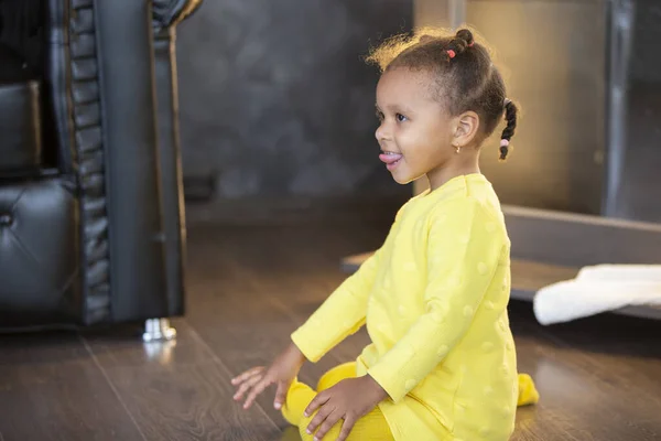 Little African American girl in a yellow dress with curly pigtails shows her tongue.