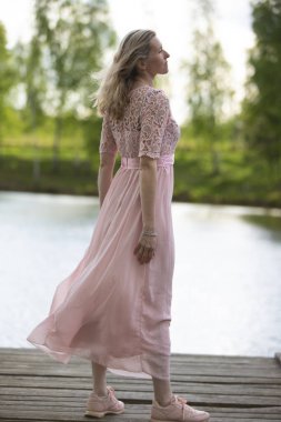 A beautiful elderly woman in a flowing pink dress stands on a wooden bridge near the river.