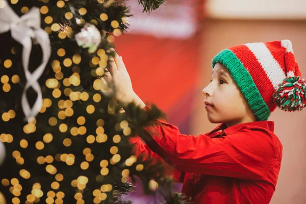 A child in a knitted Christmas hat decorates the Christmas tree.