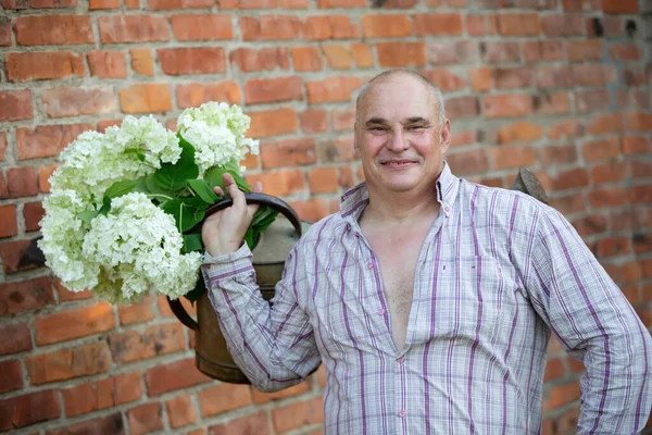 Elderly male florist. A man holds a watering can with a bouquet of flowers against a brick wall. Passion for the garden.