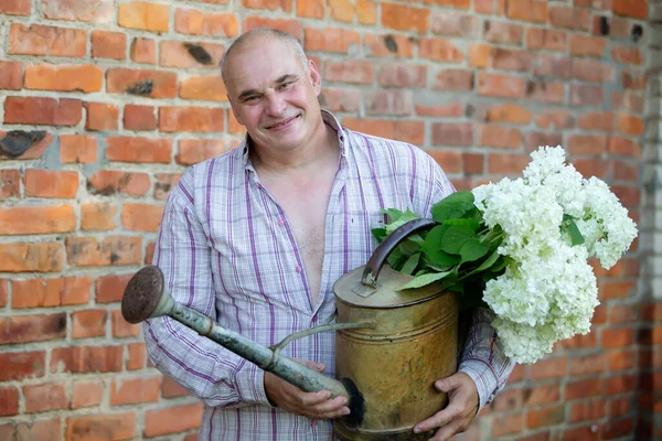 Elderly male florist. A man holds a watering can with a bouquet of flowers against a brick wall. Passion for the garden.