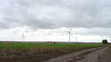 Rüzgâr parkında, arka planda bulutlu gökyüzünde büyük rüzgar türbinleri. Köy yolu. Tarladaki siluetler yel değirmenleri. Alternatif enerji.