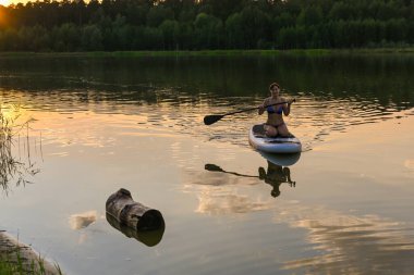 Yalnız bir kadın gün batımında çimlerle ve sık yeşil ormanlarla çevrili bir gölde Sup tahtası sürüyor. Lebyazhye Gölü, Kazan. Yaz manzarası.