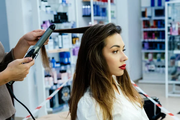 Beautiful brunette woman curls her hair with a curling iron in a beauty salon.