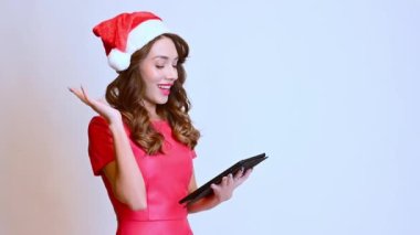 Güzel bir kadın elinde tabletle Noel şapkasıyla duruyor, beyaz arka planda gülme duyguları gösteriyor. Mutlu yıllar..