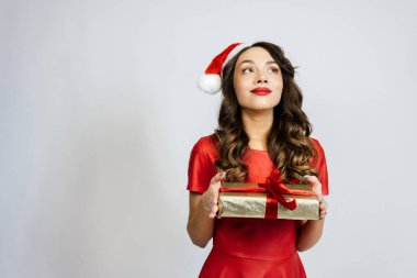 Kadın kutunun içindeki hediyeyi hayal ediyor, başka tarafa bakıyor. Beyaz bir arka planı olan Noel şapkalı güzel bir esmer..