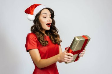 Kadın hediyeye şaşkınlıkla bakıyor. Beyaz bir arka planı olan Noel şapkalı güzel bir esmer..