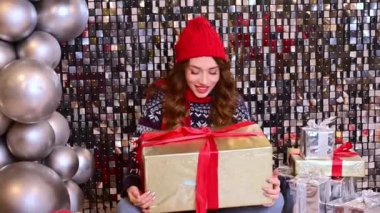 Kırmızı şapkalı güzel bir kadın, büyük bir hediyeyle eşarp yerde oturur ve büyük balonlarla ışıl ışıl bir arka planda sevinir..