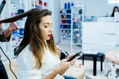 Güzel esmer bir kadın saçını güzellik salonunda şekillendirirken telefonu kullanır..