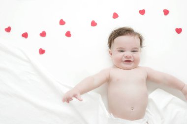 Beyaz çarşaf ve kırmızı kalp süslemeleriyle kaplı yatağında yatan beş aylık güzel ve gülümseyen bir erkek bebek. Sevgililer Günü konsepti. 