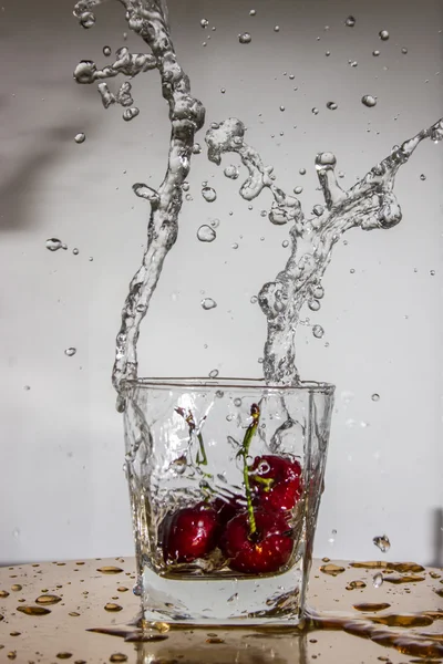 Cereja cai com um respingo em um copo com água . — Fotografia de Stock