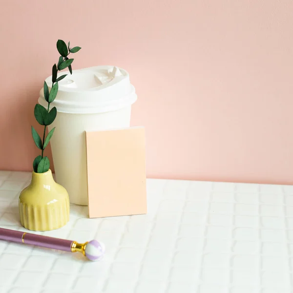 Paper coffee cup, memo pad, pen, dry plant on white desk. pink wall background