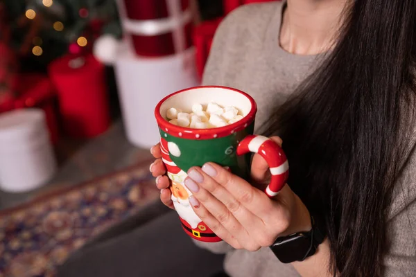 Молодая девушка в свитере держит кружку с горячим шоколадом или кофе с зефиром. Рождественские огни включены, уютная атмосфера праздника, ароматерапия для холодного зимнего сезона. рука с чашкой горячего какао — стоковое фото