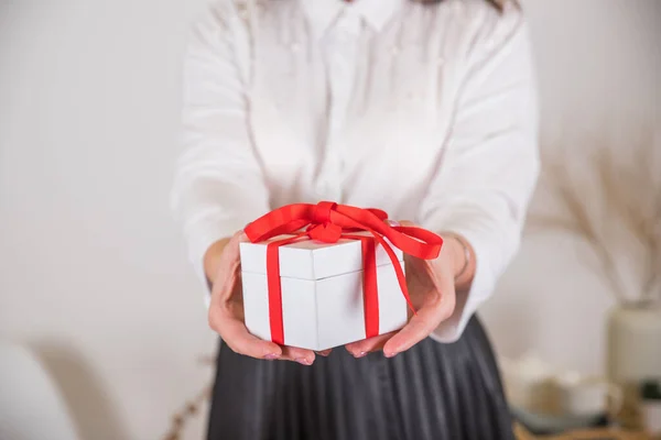 Manos femeninas sosteniendo un pequeño regalo con la costilla.Chica en camisa blanca sosteniendo una caja de regalo blanca con un lazo rojo sobre fondo borroso. Concepto de vacaciones Banner para Navidad, año de anacardo, concepto de cumpleaños. —  Fotos de Stock