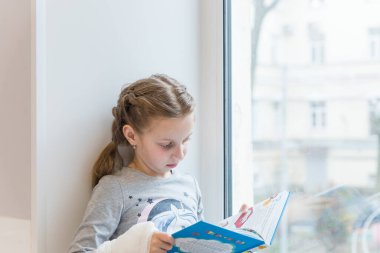 Pencere silindiri üzerine kitap okuyan sevimli küçük bir kız. Güzel, küçük Avrupalı bir kızın portresi. Konsantre olmuş görünüş, pencere kenarında dinleniyor..