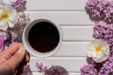 Kahve fincanı elinde. Elinde çiçekler ve hediyeyle beyaz tahta masada kahve içen genç bir kadının elleri. Bir fincan sade kahve, bahar çiçekleri, leylak çalısı. Bahar havası. Güzel kahvaltı.