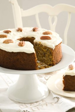 Cevizli havuçlu kek servis edilmeye hazır, bir dilim pastayla birlikte arka planda beyaz bir sandalyeyle keten masa örtüsünün üzerinde..