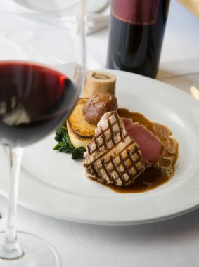 Seared Tuna steak, with beef marrow, spinach, date, potato cake and a beef and wine sauce on a white plate, complimented with a red wine.