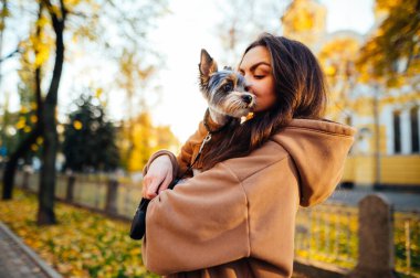 Neşeli bir köpek sahibi kadının parlak portresi. York teriyerini kucaklayan ve öpen. Sarı binanın ve ağaçların önünde duran..