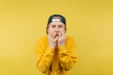 Sersemlemiş genç adam kameraya korkmuş bakıyor ve sarı arka planda çılgın bir yüzle tırnaklarını yiyor..