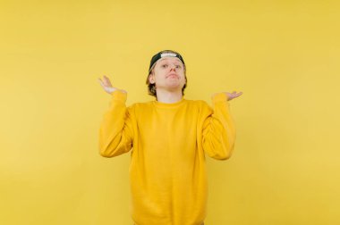 Sarı arka planda izole edilmiş, kafası karışmış genç adam kameraya bakıyor ve kollarını yana açıyor.