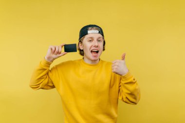 Parlak kazaklı genç adam sarı bir arka planda elinde bir banka kartıyla duruyor, kameraya bakıyor yüzünde bir gülümsemeyle ve şöyle bir jest yapıyor:.