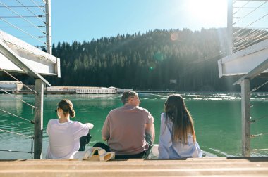 Arkadan manzara, üç arkadaş, iki kadın ve bir adam dağlarda güzel mavi bir gölün kıyısında oturmuş sohbet ediyorlar. Turistler dağlarda dinleniyor.