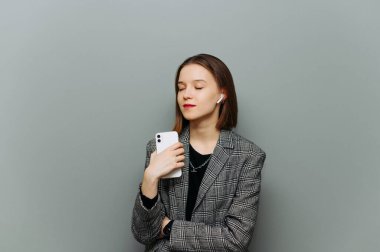 Elinde akıllı telefonuyla çekici bir kadın ve şık günlük kıyafetlerle kablosuz kulaklıkla müzik dinliyor. Gri duvarda gözleri kapalı..