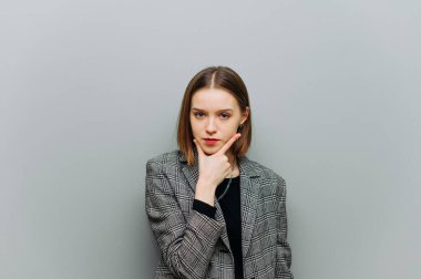 Ofis kıyafetleri içindeki düşünceli bir kadının portresi gri bir duvarın arkasında duruyor ve kameraya bakıyor ve ciddi bir yüzle düşünüyor, ceket giyiyor..