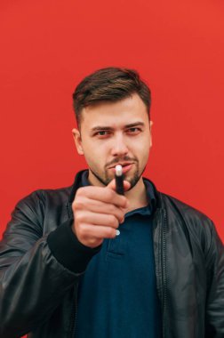 Elinde sigara tütünü olan sakallı yetişkin bir adam kırmızı bir duvarın arkasında ciddi bir yüzle kameraya bakıyor..