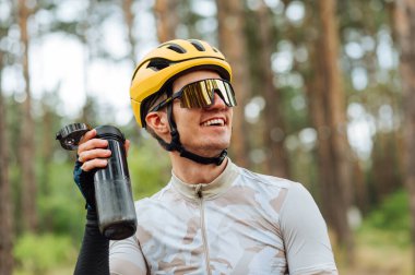 Kasklı ve gözlüklü genç bir bisikletçinin portresi. Elinde bir şişe suyla ormanın arka planında, başka yere bakıyor ve gülümsüyor..