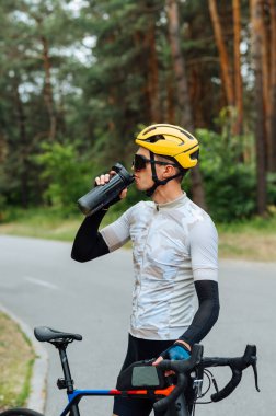 Atletik genç bisikletçi, bisiklete bindikten sonra barakadan su içer, bir orman manzarasının arka planında durur ve dinlenir ve kafasını çevirir.