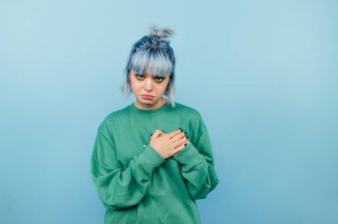 Üzgün, renkli saçlı ve makyajlı bir kız üzgün bir yüzle mavi bir arka planda duruyor ve ellerini kalbinde gezdiriyor ve kameraya bakıyor..