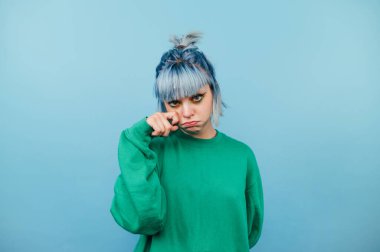 Boyanmış turkuaz saçlı üzgün hippi kız mavi arka planda durup kameraya bakarken bir gözyaşı jesti gösteriyor..