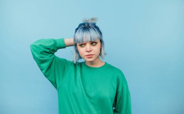 Renkli günlük giysiler ve mavi saçlar içinde güzel bir kadın mavi arka planda duruyor ve fotokopi uzayında şüpheli bir şekilde duruyor.