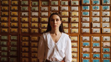 Beyaz bluzlu şık bir kadının portresi kütüphanede duruyor eski kitap arşivinin arka planında ve kameraya ciddi bir yüzle poz veriyor..