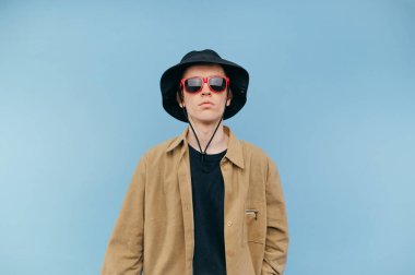 Güneş gözlüklü ve panamalı komik adam mavi arka planda izole edilmiş, ciddi bir yüzle kameraya bakıyor..