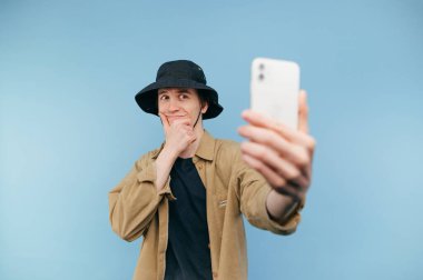 Panama 'daki genç adam mavi arka plandaki akıllı telefon kamerasına bakıyor ve selfie çekerken gülümsüyor..