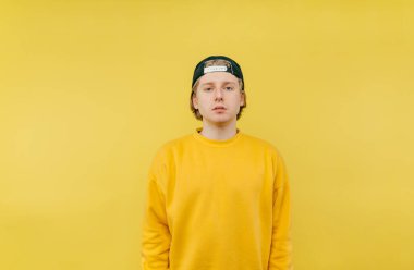 Sarı arka planda izole edilmiş şapkalı ve renkli tişörtlü yakışıklı genç adam ve ciddi bir yüzle kameraya bakıyor.