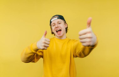 Sarı tişörtlü olumlu bir adam sarı arka planda duruyor ve parmaklarını kameraya bakarken yüzünde bir gülümsemeyle gösteriyor..