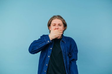 Tişörtlü uykulu adam, mavi arka planda dikilip, yorgun bir şekilde kameraya bakıyor..