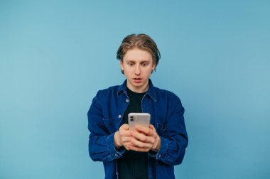 Tişörtlü, elinde akıllı bir telefonla mavi bir arka planda duran ve şok olmuş bir yüzle ekrana bakan şaşırmış genç bir adam..