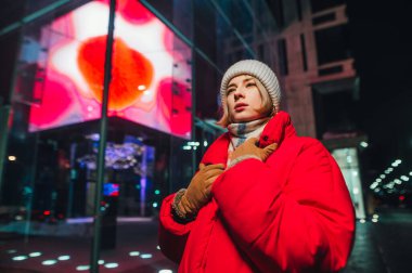 Kırmızı ceketli güzel bir kadın kış mevsiminde gece şehrinin arka planında durur ve başka tarafa bakar..