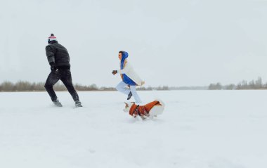 Neşeli çift karlı gölde köpekle eğleniyor, koşuyor ve gülümsüyor. Erkek ve kadın kışın karlı arazide Jack Russell 'la eğleniyor.