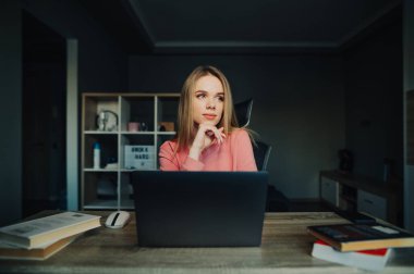 Düşünceli, çekici bir kız öğrenci dizüstü bilgisayarı ve kitaplarıyla masada oturuyor ve başka yere bakıyor ve düşünüyor..