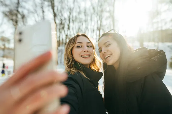 在冬天的森林里 两个漂亮而快乐的女孩在智能手机上自私自利 脸上挂着笑容 最好的朋友一起在公园里散步时被拍照 — 图库照片