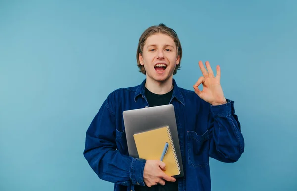 ノートパソコンとノートパソコンを手にした青い背景のシャツに身を包み 笑顔でカメラを見ている生徒の姿は Okのジェスチャーを示している — ストック写真