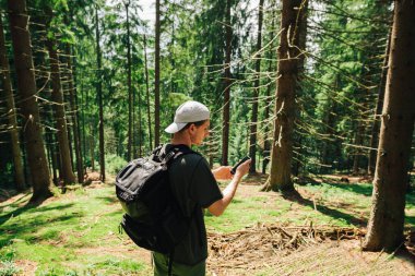 Sırtında sırt çantası olan genç bir turist dağ kozalaklı bir ormanın ortasında akıllı bir telefonla interneti kullanıyor..